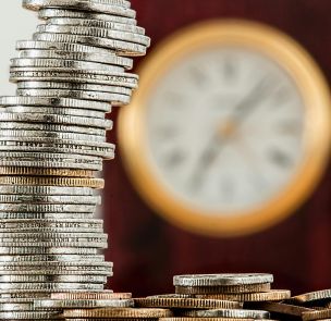 A close-up image of stacked coins with a blurred clock, symbolizing time and money relationship.